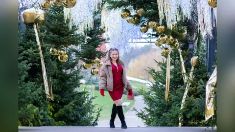 Ada — Smiling woman in a red dress and black boots posing by festive gold Christmas ornaments and evergreen trees.