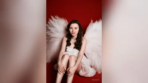 KeiraAce — KeiraAce sits with white angel wings against a red background wearing a white dress.