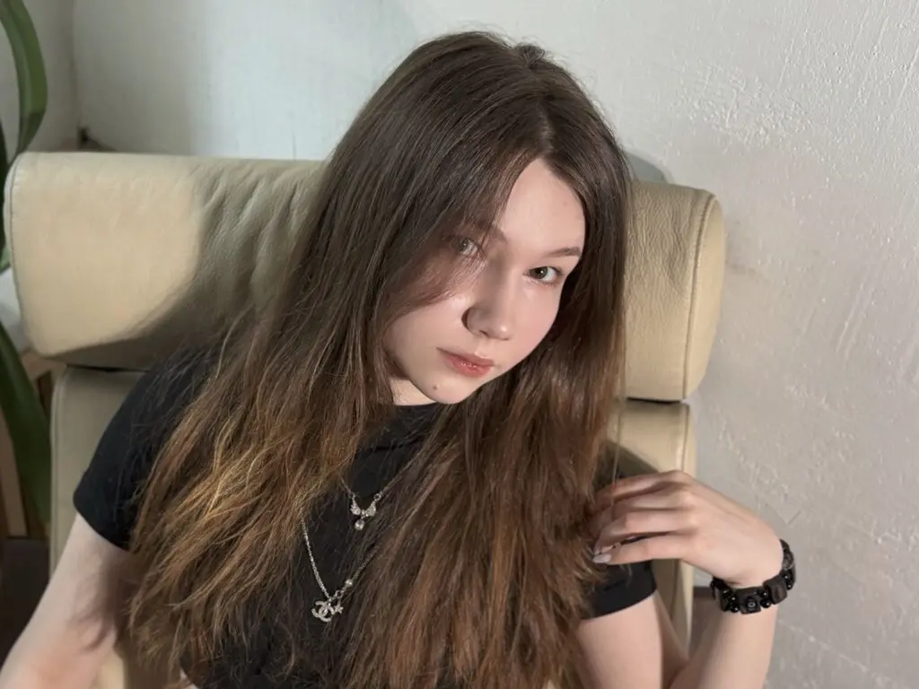 Young woman with long brown hair wearing a black t-shirt and silver necklaces sitting on a beige sofa.
