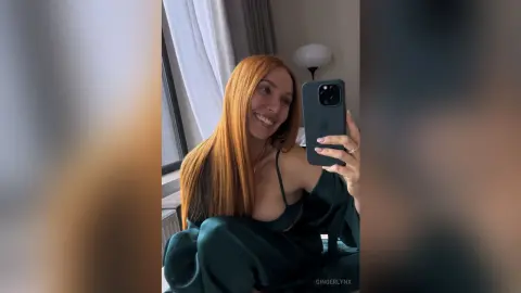 Smiling redhead woman in a green silk robe taking a selfie in a bright, airy bedroom.