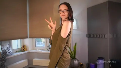 Smiling woman in an olive green cold-shoulder top making a peace sign in a bright modern living room.