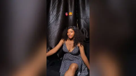 Smiling young woman in a black patterned sundress sitting against a dark curtain backdrop.