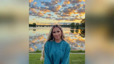 Blonde woman in a teal sweatshirt smiling by a lake during a golden sunset with dramatic clouds.
