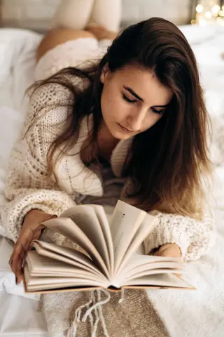 Young woman in a cozy white knit sweater reading a book on a bed in a relaxing, peaceful atmosphere.