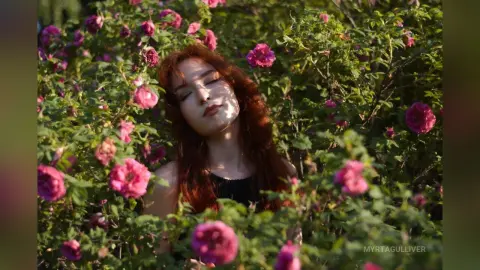 Redheaded woman in a black top resting peacefully amidst blooming pink rose bushes in a sunny garden.