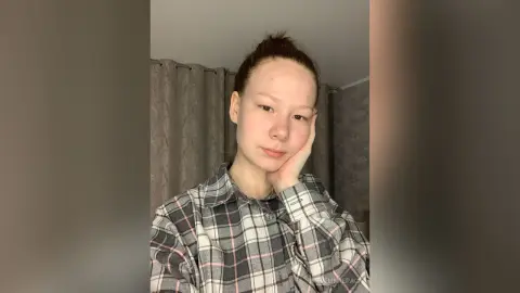Young woman in a grey plaid flannel shirt posing thoughtfully in a room with grey curtains.