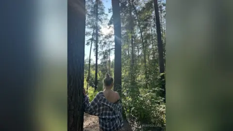 Woman in a plaid shirt leaning against a tree in a sunlit forest setting with a peaceful mood.