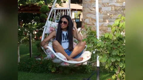 Young woman in a blue tank top and sunglasses relaxing in a hanging swing chair in a lush garden setting.