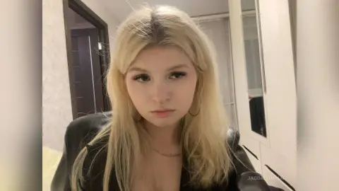 Blonde woman in a black leather jacket posing with a moody expression in a modern minimalist room.