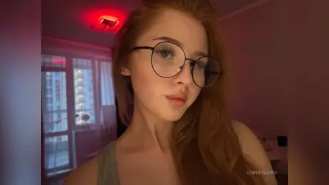 Redheaded woman with round glasses in a moody, red-lit room wearing a grey tank top.