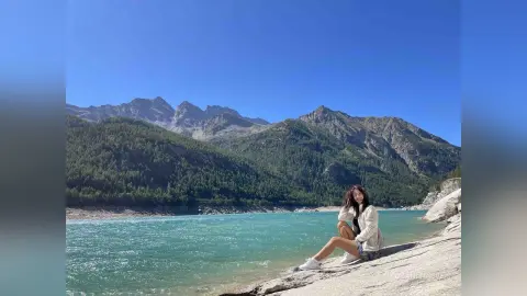 DyandraEve sits on a rocky shore by a blue lake, wearing a white top and denim shorts.