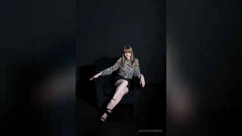 NatashaSunrise — NatashaSunrise sits on a dark armchair wearing a black and white striped shirt and a short black skirt.