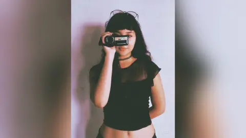 Young woman with dark hair and black top holding a vintage video camera against a plain wall.