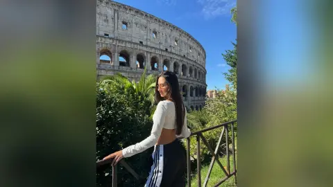 AlanaPearce poses outdoors in a white long-sleeve crop top and black athletic pants with white stripes, standing by a railing with the Colosseum in the background.