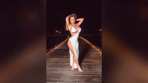 Blonde woman in a white cut-out maxi dress posing on a wooden pier at night under warm lighting.