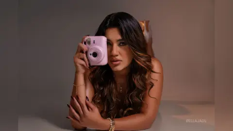 Glamorous woman in gold jewelry taking a mirror selfie with a pink phone in a moody studio setting.