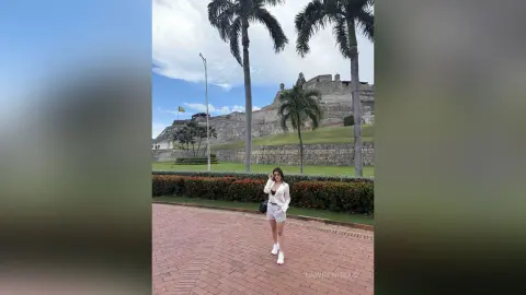 Woman in white blouse and shorts standing by a stone wall and palm trees at a historic castle site.