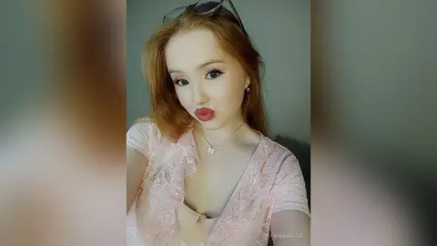 Young woman with long blonde hair wearing a pink lace top and necklace posing with a playful expression.
