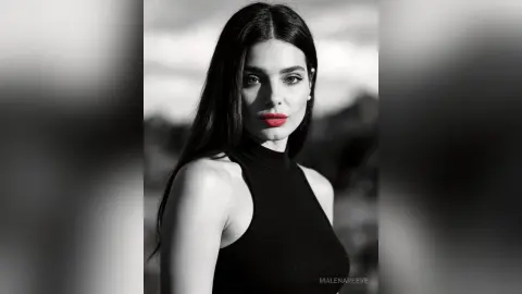 Elegant woman with long dark hair and red lips wearing a black sleeveless turtleneck in a moody outdoor setting.