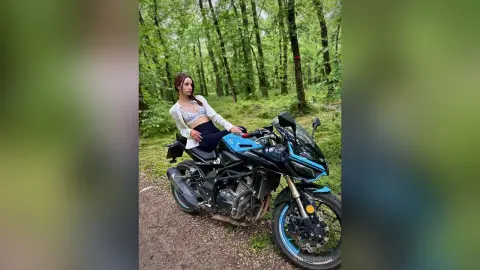 Stylish woman in a white crop top sitting on a black sport motorcycle in a lush green forest setting.