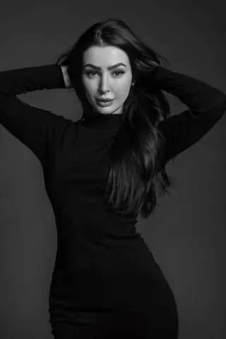 Anelinsa poses with her hands in her long dark hair, wearing a tight black long-sleeve dress against a dark background.