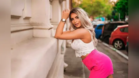 Smiling blonde woman in a white lace crop top and pink shorts posing against a stone wall in a sunny street.