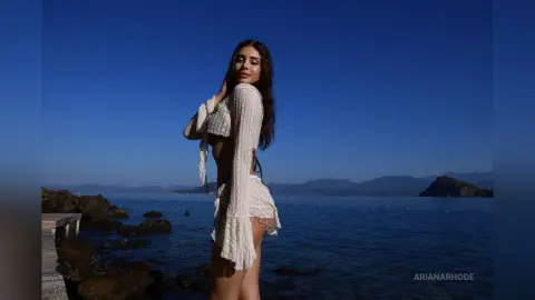 Young woman in a white lace boho crop top and shorts posing by the ocean under a clear blue sky.