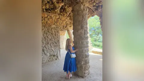 Blonde woman in a blue summer dress standing by ancient stone ruins in a sunny outdoor setting.