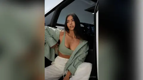 Stylish young woman in a sage green jacket and crop top posing confidently by a white vehicle.