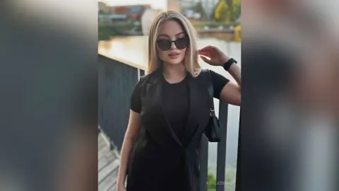AshlieBridge wears a black sleeveless top and dark sunglasses while standing outdoors near a wooden deck.