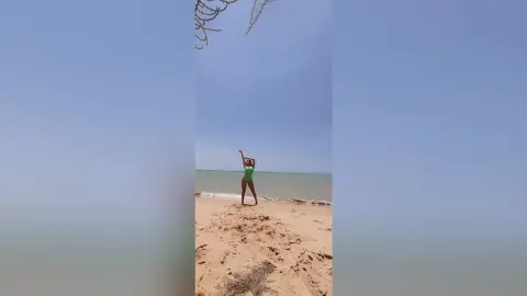 Cheerful woman in a green swimsuit standing on a sunny sandy beach under a clear blue sky.