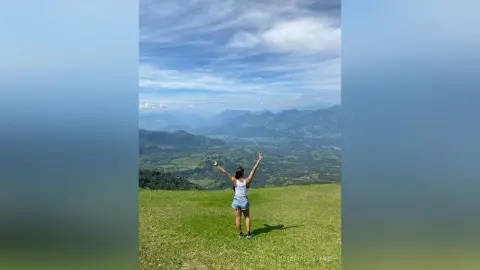 VictoriaFontaine stands in a grassy field with her arms raised high, overlooking a vast mountain valley under a cloudy blue sky.