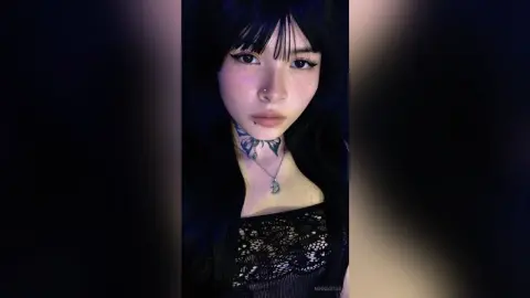 Young woman with dark hair and neck tattoo wearing a black lace top in moody, low-light aesthetic.