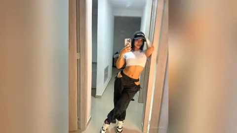 Young woman in white crop top and black cargo pants taking a mirror selfie in a modern minimalist room.