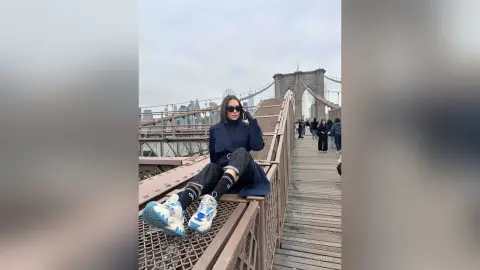 Stylish woman in dark coat and sunglasses sitting on Brooklyn Bridge walkway against city skyline.