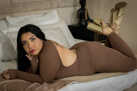 Curvy woman in a brown ribbed bodysuit relaxing on a bed in a cozy bedroom setting.