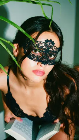 Mysterious woman in a black lace mask and lingerie reading a book in a moody, leafy indoor setting.