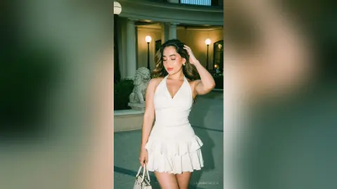 LilianMendoza poses outdoors in a white ruffled romper, running her hand through her long wavy hair while holding a small white handbag.