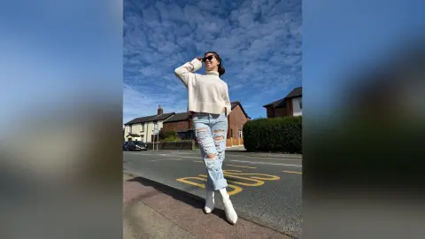 Stylish woman in a white sweater and ripped jeans posing on a sunny street under a blue sky.