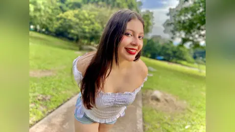 Smiling young woman in a white off-the-shoulder top and denim shorts walking on a sunny park path.
