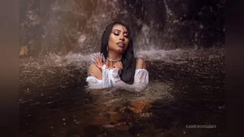 Serene woman in a white off-the-shoulder dress relaxing in a dark forest waterfall pool.