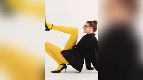 Fashionable woman in bright yellow tights and black blazer posing playfully against a white studio background.