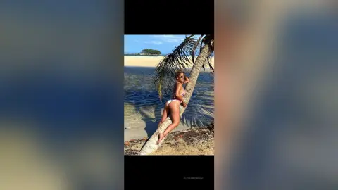 AlexiaMendoza poses in a white bikini while leaning against a palm tree on a tropical beach.