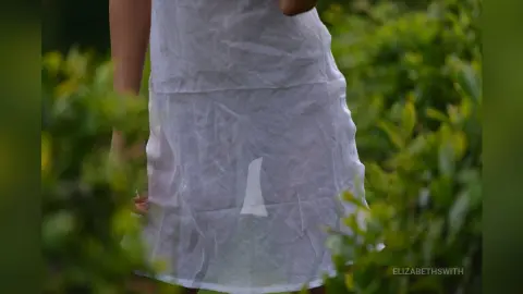 Woman in a breezy white linen dress standing in a lush green garden setting.