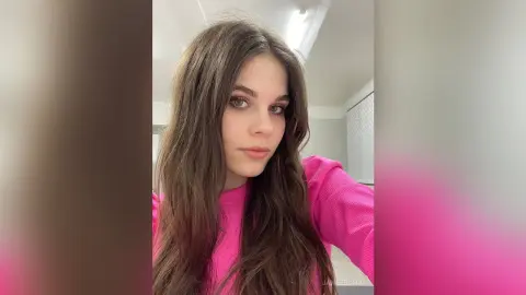 Young woman with long brown hair wearing a pink ribbed top in a bright indoor setting with a calm expression.