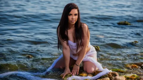 Brunette woman in a white flowing dress kneeling on rocks by the blue ocean under bright sunlight.