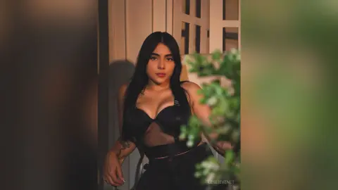 Young woman in a black mesh bodysuit posing in warm, moody indoor lighting.