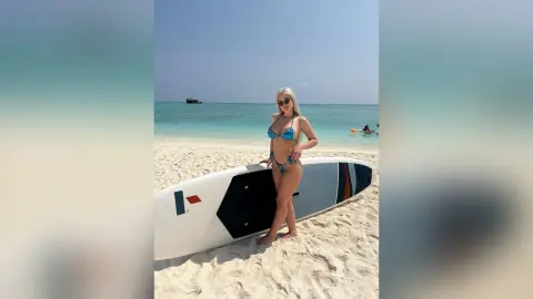 Blonde woman in a blue bikini standing by a white surfboard on a sunny tropical beach.