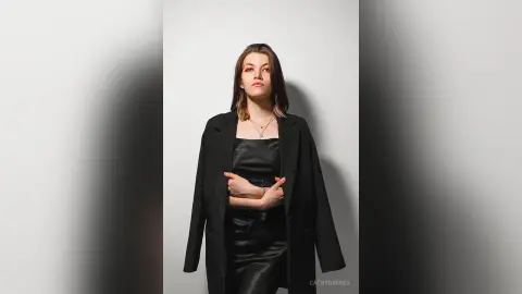 Elegant woman in a black satin dress and overcoat posing against a minimalist white background.