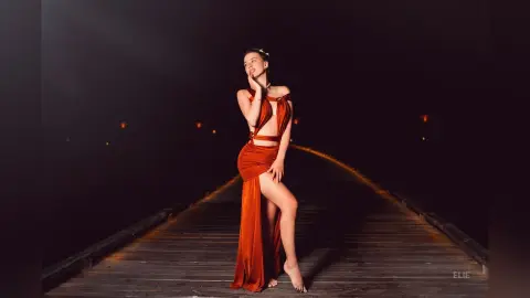 Elie poses in a long, red cut-out dress with a high slit, standing on a wooden walkway against a dark background.